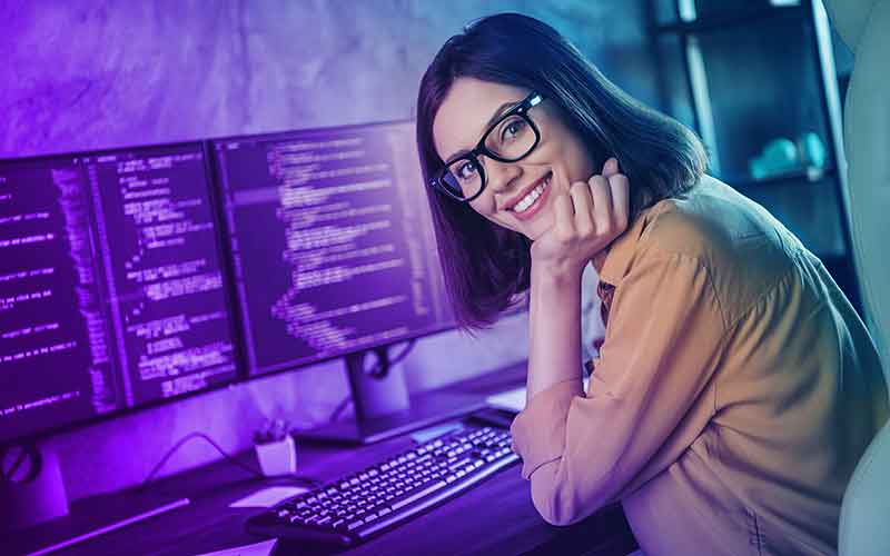 Junge Frau sitzt an einem Arbeitsplatz mit drei Monitoren voller Code – motivierte IT-Studentin bei Printus