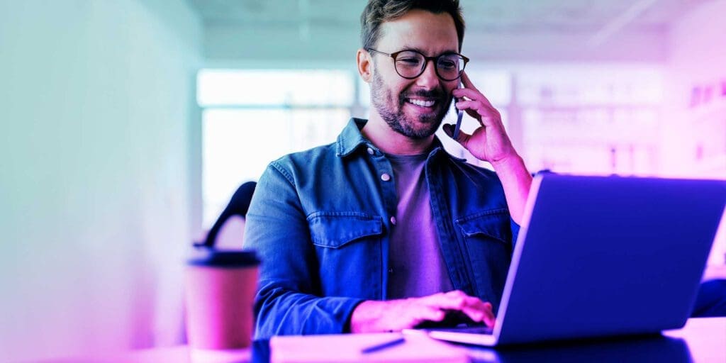 Lächelnder Mann mit Brille telefoniert und arbeitet am Laptop im Büro