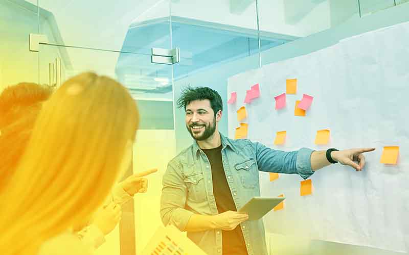 Junger Mann präsentiert vor einem Whiteboard mit bunten Post-its – interaktive Teamarbeit und kreative Projektarbeit bei Printus