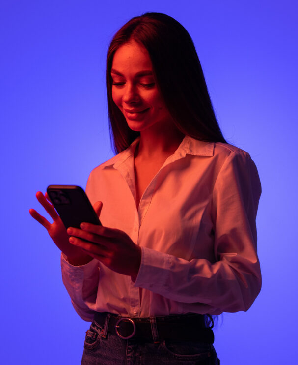 Eine junge Frau in weißer Bluse blickt mit einem Lächeln auf ihr Smartphone. Der Hintergrund ist in kräftigem Blau- und Rottönen gehalten. Sie wirkt organisiert, souverän und modern.