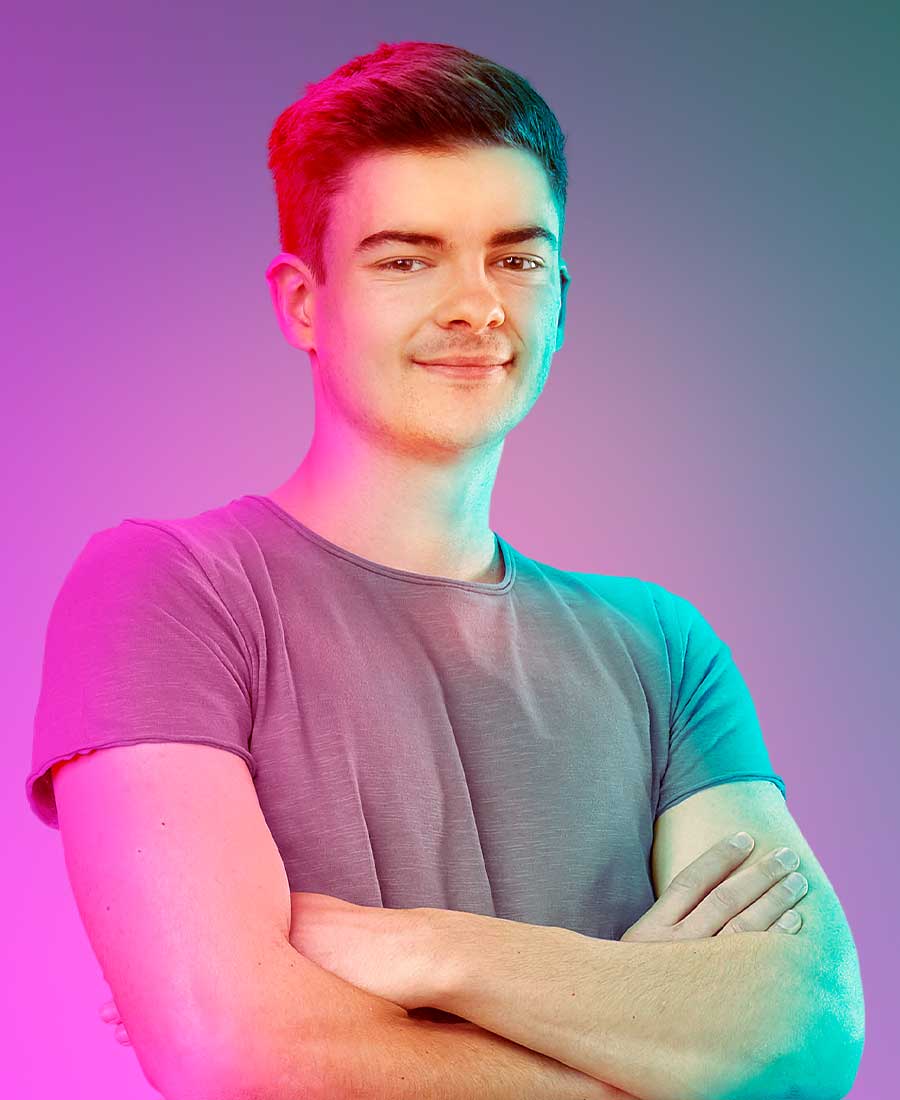Junger Mann mit kurzen dunklen Haaren, grauem T-Shirt und verschränkten Armen schaut selbstbewusst in die Kamera. Der Hintergrund ist bunt beleuchtet in Pink-, Blau- und Grüntönen.