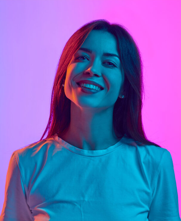 Eine junge Frau mit dunklen Haaren und hellem Shirt lächelt freundlich in die Kamera. Das Bild ist in kontrastreiche Blau- und Pinktöne getaucht und symbolisiert die Verbindung von technischer Expertise und kreativer Marketingumgebung im Bereich DevOps und Systemadministration.