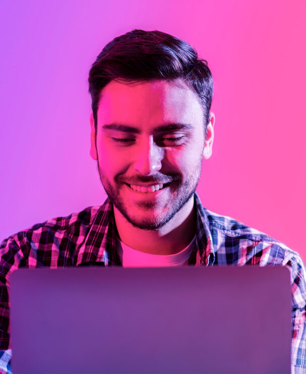 Junger Mann mit kariertem Hemd arbeitet lächelnd am Laptop. Die pink-violette Beleuchtung verleiht dem Bild einen modernen Look.