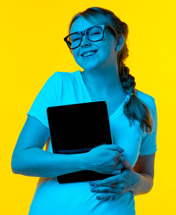 Eine junge Frau mit Brille und geflochtenem Zopf steht vor einem gelben Hintergrund, lächelt selbstbewusst und hält ein Tablet im Arm. Sie verkörpert den idealen Mix aus Technikaffinität und Teamspirit.