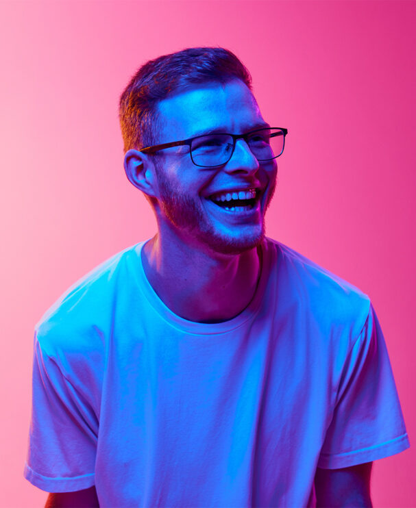Ein junger Mann mit Brille und Bart lacht herzlich und trägt ein weißes T-Shirt. Die bunte Beleuchtung in Pink und Blau erzeugt eine energiegeladene, zukunftsorientierte Atmosphäre.