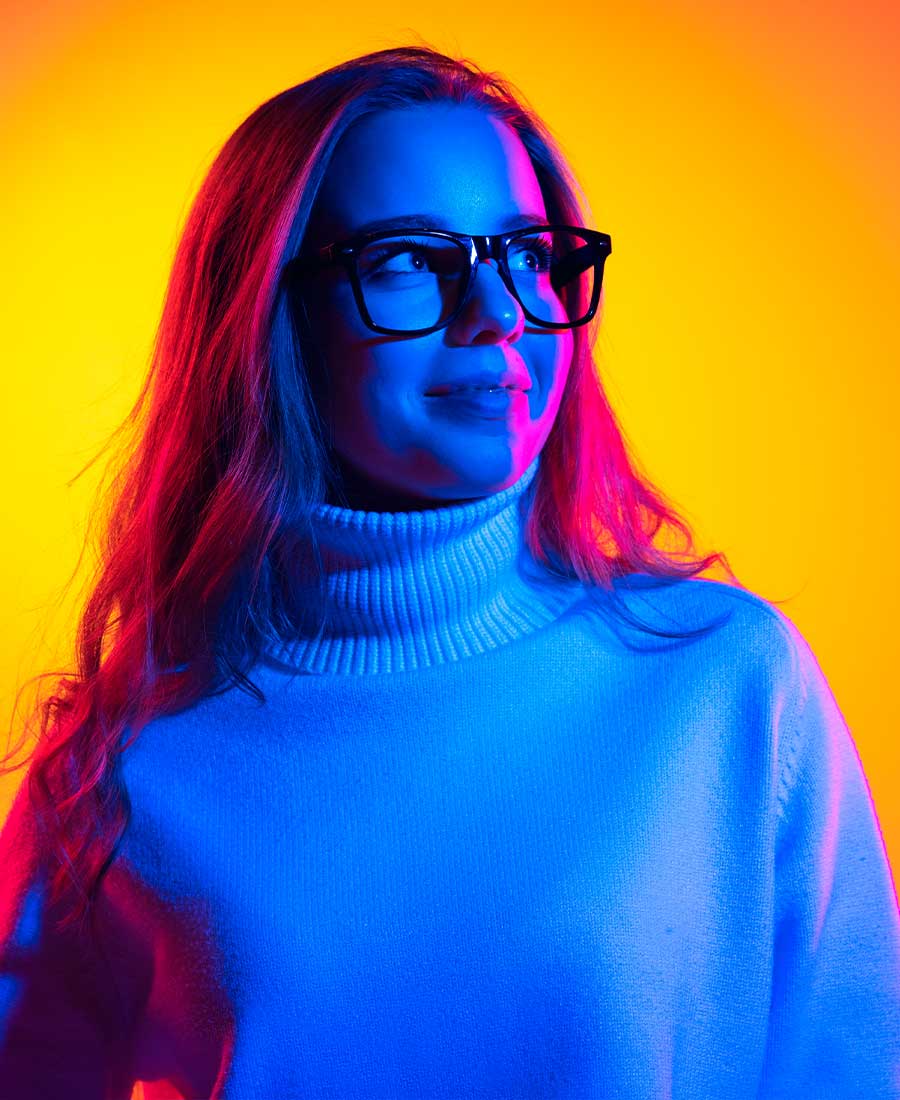 Eine junge Frau mit langen Haaren, Brille und hellem Rollkragenpullover blickt lächelnd nach oben. Sie ist in leuchtendes gelb-orangefarbenes und blau-violettes Licht getaucht, was eine moderne und zukunftsorientierte Stimmung vermittelt.