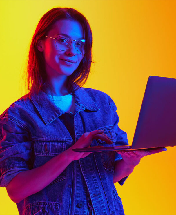 Junge Frau steht mit dem Laptop in der Hand vor einem gelben Hintergrund