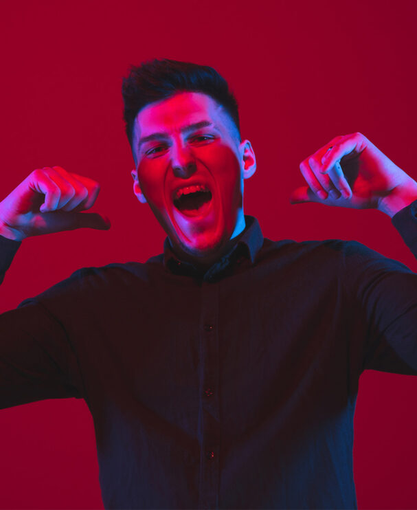 Ein junger Mann in dunklem Hemd jubelt lautstark und zeigt mit beiden Daumen auf sich selbst. Die intensive Beleuchtung in Rot und Blau erzeugt eine energiegeladene, selbstbewusste Stimmung.