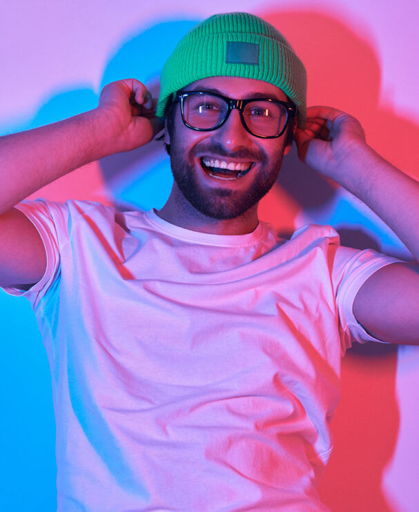 Ein junger Mann mit Brille, grünem Beanie und weißem T-Shirt lacht herzlich und fasst sich an die Mütze. Die farbige Ausleuchtung in Blau und Magenta erzeugt eine lebendige, moderne Stimmung.
