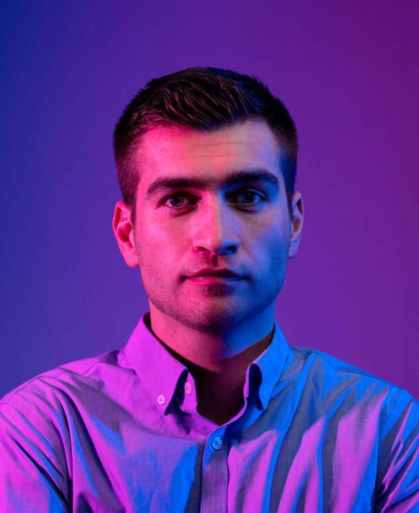 Ein junger Mann in einem hellen Hemd blickt ruhig und fokussiert in die Kamera. Die Beleuchtung in kräftigem Blau und Violett schafft eine technische, moderne Atmosphäre und unterstreicht seine Rolle als Maschinen- und Anlagenführer im logistischen Umfeld.