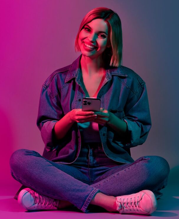 Eine junge Frau im Jeans-Outfit sitzt entspannt im Schneidersitz auf dem Boden und hält lächelnd ein Smartphone in der Hand. Die Beleuchtung in Pink und Blau vermittelt eine moderne, digitale Atmosphäre.