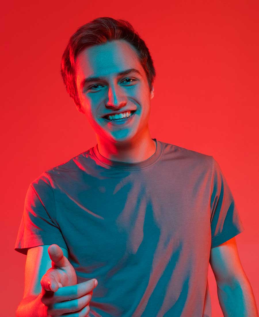 Ein junger Mann in einem schlichten T-Shirt lächelt offen in die Kamera und zeigt mit einer einladenden Geste nach vorn. Die Ausleuchtung in Rot und Blau vermittelt Energie, Offenheit und Zukunftsorientierung.