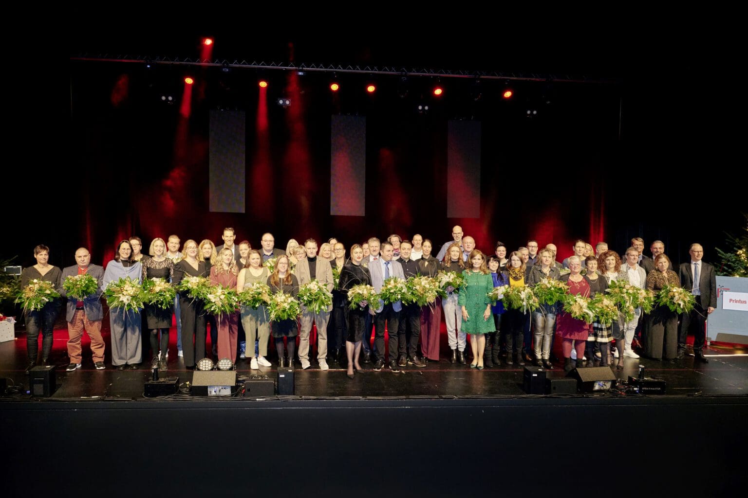 Gruppenfoto der Printus-Jubilare bei feierlicher Ehrung auf der Bühne, mit Blumensträußen und rotem Bühnenlicht