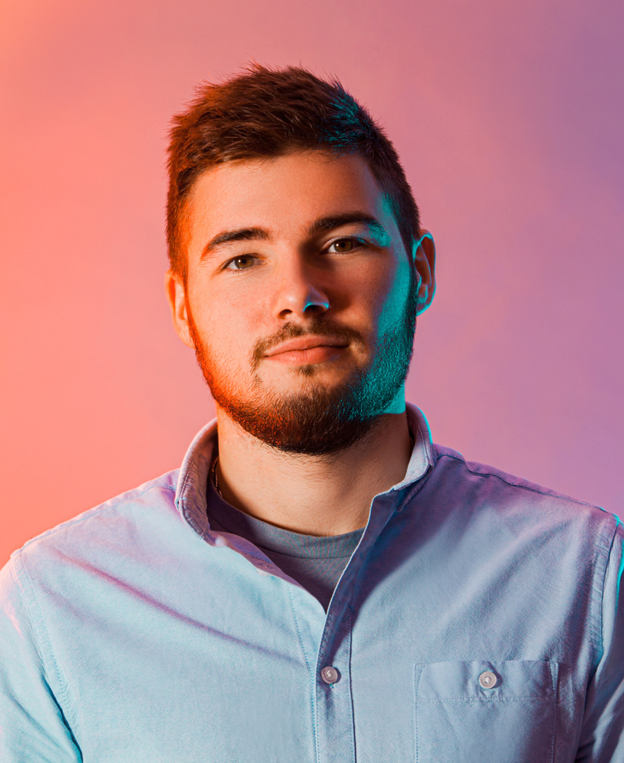 Junger Mann mit kurzem Haar und Bart blickt selbstbewusst in die Kamera. Er trägt ein helles Hemd, der Hintergrund ist in warmen Orange- und Rottönen gehalten.