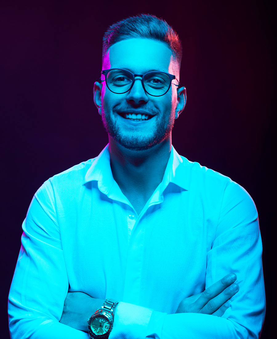 Junger selbstbewusster Mann mit Blick in die Kamera, leicht lächelnd, mit neonfarbener Beleuchtung auf dunklen Hintergrund.